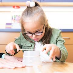 A girl sits with a teacher at a desk working on an upturned plastic container for the Bye Bye Bug activity