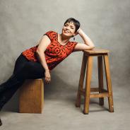 Mexican woman with short dark hair. Sitting on a wooden bench and leaning to her side with her elbow on a stool. Leslie is wearing black pants and a short-sleeved red top.