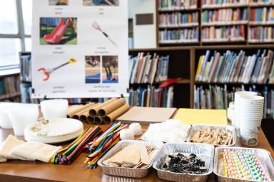 A library with a table full of classroom engineering design supplies: straws, paper, clips, pipecleaners, paper plates, tape and glue
